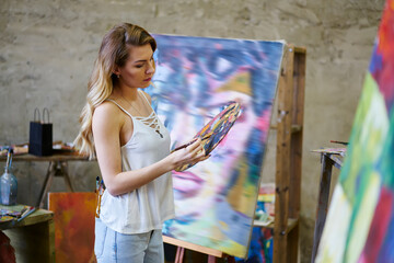 Young female artist mixing acrylic colors during hobby time in loft studio for art workshop relaxing, Caucasian woman dressed in casual clothes holding palette checking details with paintbrush