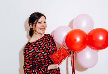 Valentine's Day. Portrait of a beautiful young woman in red dress holding red balloons and red gift box on white background. Be my Valentine