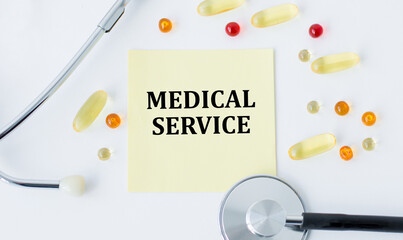 Wall Mural - Medical concept, the inscription - MEDICAL SERVICE on a yellow sticker on a white background around the scattered tablets and lies a stethoscope