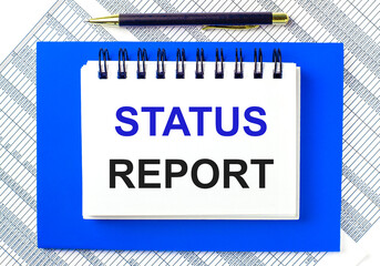 Fototapete - Against the background of reports on the desktop, a blue notepad. It has a pen and a white notebook with the text STATUS REPORT. Business concept