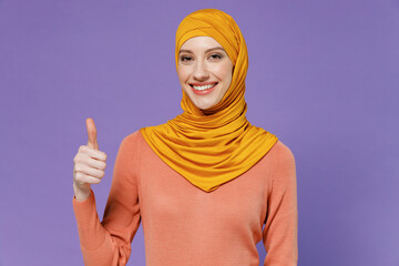 Wall Mural - Smiling jubilant overjoyed excited happy young arabian asian muslim woman in abaya hijab yellow clothes showing thumb up like gesture isolated on plain pastel light violet background studio portrait.