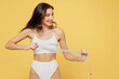 © ViDi Studio - Lovely sexy smiling young brunette woman 20s wear white underwear with perfect fit body standing posing hold measure tape on breast look aside on copy space area isolated on plain yellow background.