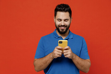 Wall Mural - Young smiling fun cool happy cheerful man 20s wear basic blue t-shirt hold in hand use mobile cell phone browsing chatting isolated on plain orange background studio portrait People lifestyle concept