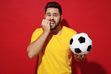 Wall Mural - Worried vivid young bearded man football fan in yellow t-shirt cheer up support favorite team hold in hand soccer ball look camera biting nails isolated on plain dark red background studio portrait.