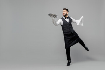 Wall Mural - Full body young smiling barista male waiter butler man wear white shirt vest elegant uniform work at cafe carrying walk give metal tray isolated on plain grey background Restaurant employee concept.