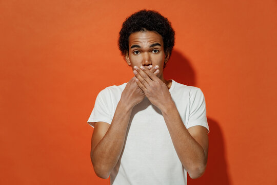 Frightened scared sad shocked panicked pop-eyed startled amazed young black curly man 20s years old wears white t-shirt over mouth with hand isolated on plain pastel orange background studio portrait.
