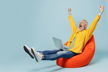 Full Size Excited Elderly Gray-haired Mustache Bearded Man 50s Wear Yellow Shirt Sit In Bag Chair Hold Use Work On Laptop Pc Computer Spread Hands Isolated On Plain Pastel Light Blue Background Studio