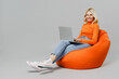 © ViDi Studio - Full body elderly smiling happy blonde woman 50s in orange turtleneck sit in bag chair hold use work on laptop pc computer isolated on plain grey background studio portrait. People lifestyle concept.