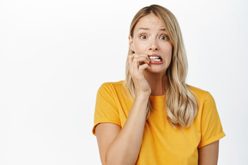 Wall Mural - Nervous blond girl, anxious young woman biting fingernails, looking guilty or worried, standing against white background