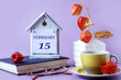 Calendar for February 15: the name of the month in English, the numbers 15, a yellow cup of tea, a physalis branch in a vase, a book, glasses on a pastel background
