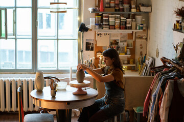 Poster - Creative young woman focused on pottery making in small studio. Female artist spend time working with clay shaping handcraft tableware for selling in handmade potter store or workshop artistic space