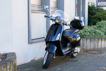 A Black Scooter With A Luggage Rack Is Parked Against The Building With Windows.