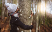 Middle-aged Woman In White Cap Hugs And Kisses A Tree Trunk At Sunset. Concept Connection With Nature. Balneotherapy.