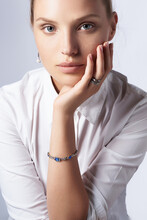Close-up Shot Of A Blonde European Lady In A White Shirt, Wearing An Earring, Rings And A Rhinestone Bracelet With Blue Crystals Shaped As Hearts. The Photo Is Made On The Gray Background. 