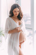 Young beautiful pregnant woman stand near window hugs bare belly with hands. White long silk negligee pregnancy dress peignoir, dressing gown. Light room classic background. Gently smiling face