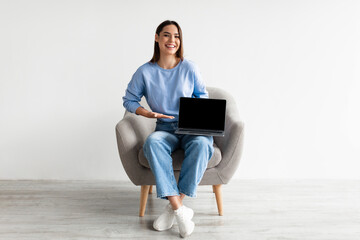Poster - Happy young lady sitting in armchair with laptop, showing blank screen with mockup, advertising your website design