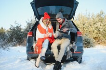 Couple in love sitting in car trunk drinking hot tea in snowy winter forest and chatting. People relaxing outdoors during road trip. Valentines day