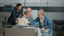 Senior Man Receiving Visit From Mother And Little Girl In Hospital Ward. Child With Flowers Visiting Grandpa With Illness In Bed. Elder Patient Enjoying Chat With Family And Visitors At Clinic.