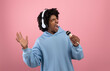 © Prostock-studio - Creative Afro teen in headphones listening to music, singing karaoke song into microphone on pink background