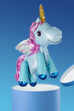 Shot Of A Balloon Shaped As A Light Blue Unicorn. It Has A Golden Horn, A Pink Tail And A Mane, Multicolored Wings. The Multicolored Balloon Is Flying Out Of A Box On A Blue Background.