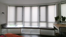 Video of motorized roller shades in the interior. Automatic roller blinds beige color open up on big glass windows. Curtaines are above the windowsill with pillows. Summer. Green trees outside.