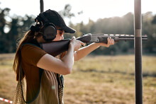 Young caucasian female on tactical gun training classes. Woman with weapon, wearing cap, protective headphones and eyeglasses. Outdoor Shooting Range. At Sunny evening at summer. Rear view