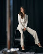 © Dmitry Tsvetkov - Long haired businesswoman in elegant formal white pantsuit with high heeled shoes sits on bar stool on black background in studio