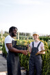© Roman - team of researchers at work in greenhouse, female with notepad and pen in hand, stand talking with black guy in uniform. Greenhouse production. Female farm worker examine and checking plants