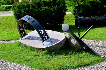 A close up on a sun dial and sun clock made out of metal and stone, located next to an old abandoned anchor of a big ship located in the middle of a dense public park next to a gravel path or road