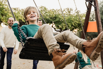 LGBT family - Happy children sons and fathers having fun swinging on swing at city park - Focus on left kid face