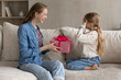 © fizkes - Joyful little girl sitting on couch with covered eyes, feeling excited waiting for wished item in wrapped box. Happy caring young mother congratulating small daughter with birthday, preparing gift.