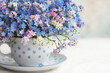 Bouquet of spring forget-me-not flowers in a cup on the table against the background of a colored wall. Holiday card, fragment, blur, selective focus.