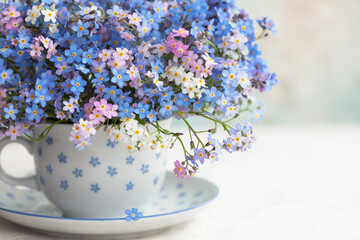 Wall Mural - Bouquet of spring forget-me-not flowers in a cup on the table against the background of a colored wall. Holiday card, fragment, blur, selective focus.