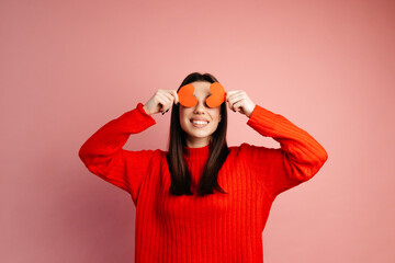 Wall Mural - Cute girl is smiling and covering her face with paper hearts. The girl is standing isolated against the pink background. Concept of the St. Valentine's Day