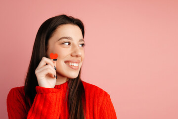 Wall Mural - Attractive girl is looking on the left and standing isolated against the pink background. The girl is holding a tiny red heart in the hand. Concept of the St. Valentine's Day