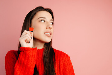 Wall Mural - Beautiful girl is looking on the left and standing isolated against the pink background. The girl is holding a tiny red heart close to her face. Concept of the St. Valentine's Day