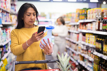 Hispanic Woman Scanning Barcode On Detergent Bottle With Her Smartphone While Shopping In Supermarket, Checking Product Information And Price. Modern Technology For Convenience Of Buyers
