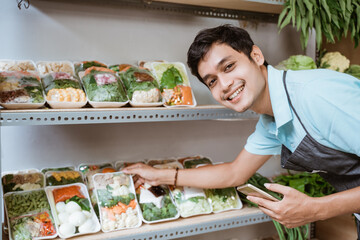 Wall Mural - Smiling man selling vegetables holding a smartphone while checking vegetables