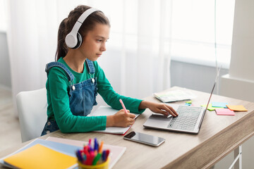 Poster - Focused girl sitting at desk, using laptop, writing in copybook