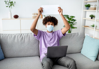 Wall Mural - African American youth in face mask showing empty sign with mockup, sitting on couch with laptop during covid