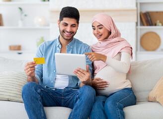 Wall Mural - Excited pregnant islamic spouses shopping online with tablet and credit card at home, relaxing together on sofa