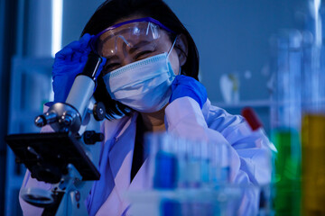 Wall Mural - Studio shot of Asian professional female middle aged scientist in white lab coat safety glasses face mask and rubber gloves using microscope lens looking at covid19 coronavirus sample in laboratory