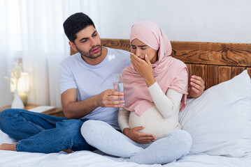 Wall Mural - Care and support concept. Arab expecting wife feeling sick, husband giving her glass of water, sitting on bed