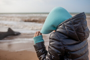 Teenager female walking outdoors in the beach in cold weather with wind, look to the sea, Girl dreased in black jacket and blue hoody sweater and trousers