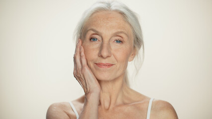 Closeup Portrait of Senior Woman Looking at Camera, Touching Hair, Beautiful Face, Smiling. Elderly Lady with Natural Grey Hair, Blue Eyes. Abstract Beige Background, Tender and Soft Moment