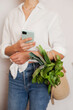 © Dina - person in white shirt and jeans holding a mobile phone and eco jute reusable bag with bunch of green basil and lettuce. Grocery shopping for vegetables - healthy lifestyle