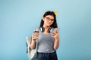 Wall Mural - Indian teen female student with backpack and headphones holding takeaway coffee, using smartphone on blue background