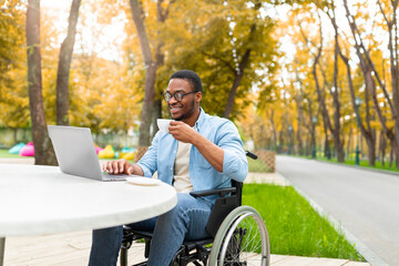 Wall Mural - Remote job for impaired people. Cheerful black guy in wheelchair working online, using laptop at outdoor cafe in autumn