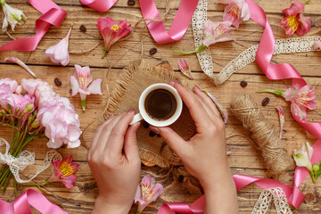 Wall Mural - Hands with a coffee cup on a wooden table between pink flowers, ribbons and coffee beans