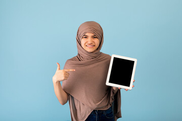 Wall Mural - Joyful teen girl in Muslim hijab pointing at tablet pc with blank screen on blue background, mockup for app or website